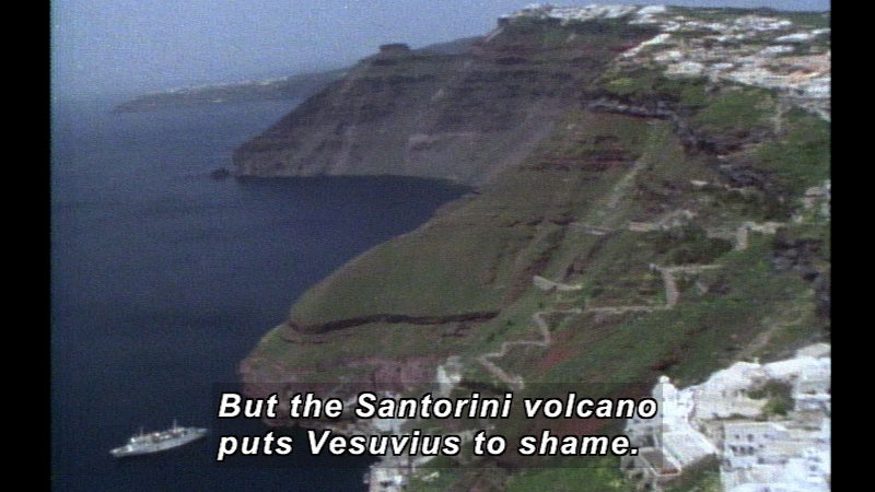 Aerial view of a steeply descending hillside that ends in water. A cruise ship in in the water at the foot of the hillside. Caption: But the Santorini volcano puts Vesuvius to shame.