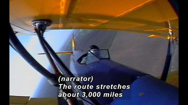 Person in the open cockpit of a small plane. Caption: (narrator) The route stretches about 3,000 miles