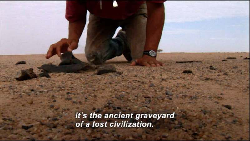 Person kneeling on the ground and reaching for an object. Caption: It's the ancient graveyard of a lost civilization.