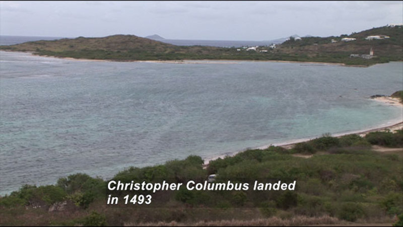 A protected bay of light green water with a handful of buildings slightly set back from the coastline. Caption: Christopher Columbus landed in 1493