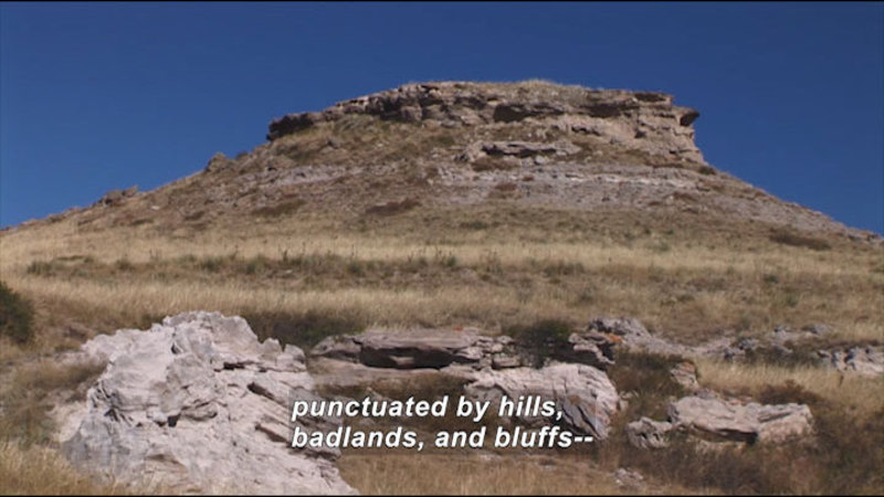 Dry, brown grass and brush with crumbling rocks throughout and a flat-topped rocky hill in the background. Caption: punctuated by hills, badlands, and bluffs --