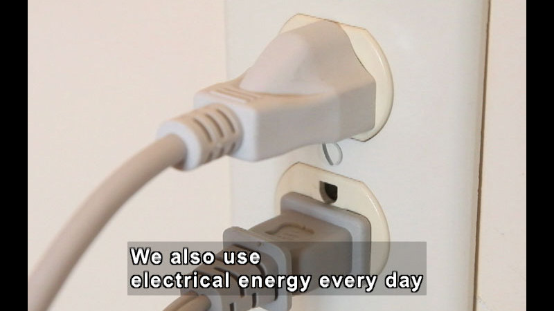Two power cords plugged into a wall outlet. Caption: We also use electrical energy every day