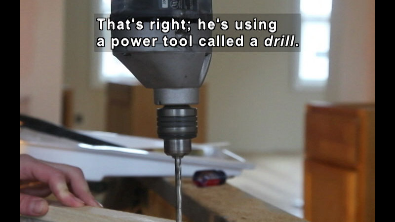 Thin tubular object protruding from a machine casing descends perpendicular to a piece of wood laying horizontally below it. Caption: That's right; he's using a power tool called a drill.