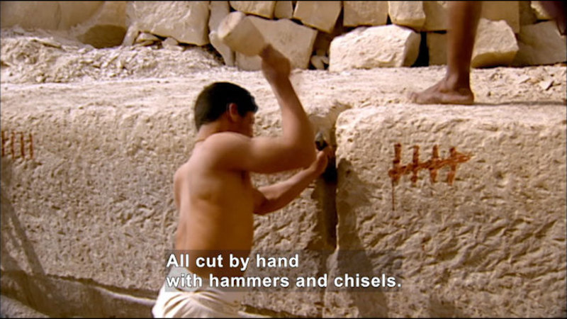 Person in a loin cloth chiseling a large block of light-colored stone. The block has hashmarks representing "5". Caption: All cut by hand with hammers and chisels.