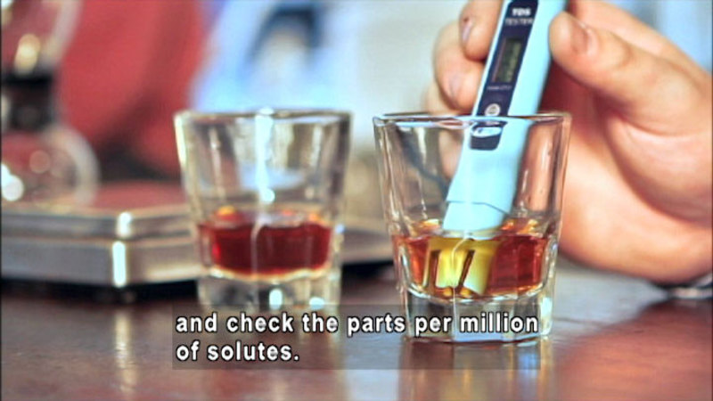 Two glasses with colored liquid in them. One glass has a person holding a cylindrical electronic device in the liquid. Caption: and check the parts per million of solutes.