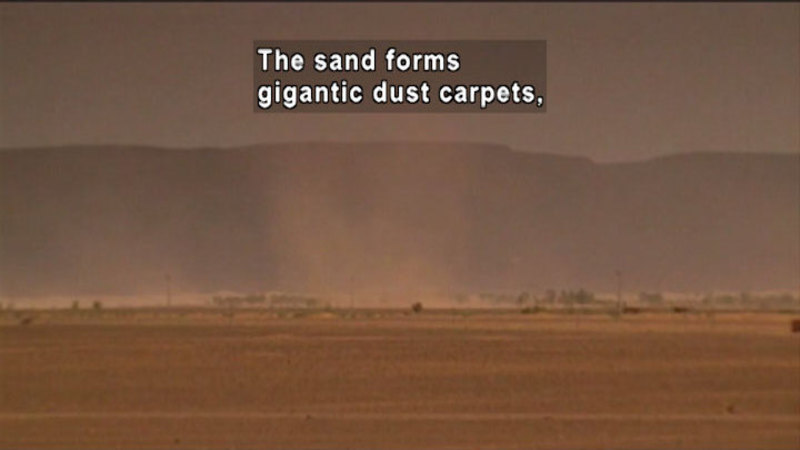 In foreground a flat, barren landscape with a large cloud of dust. In the background there are some out-of-focus hills. Caption: The sand forms gigantic dust carpets,