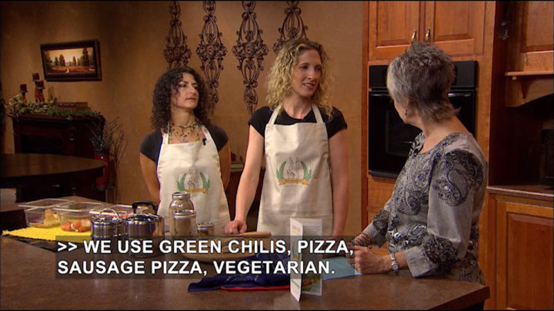A woman talking to two other women in aprons. On the counter are cooking supplies. Caption: We use green chilis, pizza, sausage pizza, vegetarian.