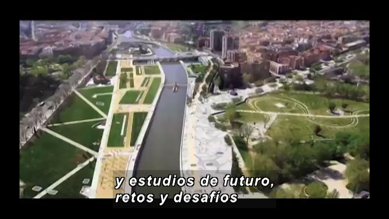 A channel of water blanketed by green spaces cuts its way through a big city. Spanish captions.