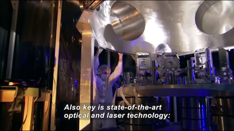Person in lab coat, gloves, and mask moving a large metal machine that is place above other complex metal machinery. Caption: Also key is state-of-the-art optical and laser technology: