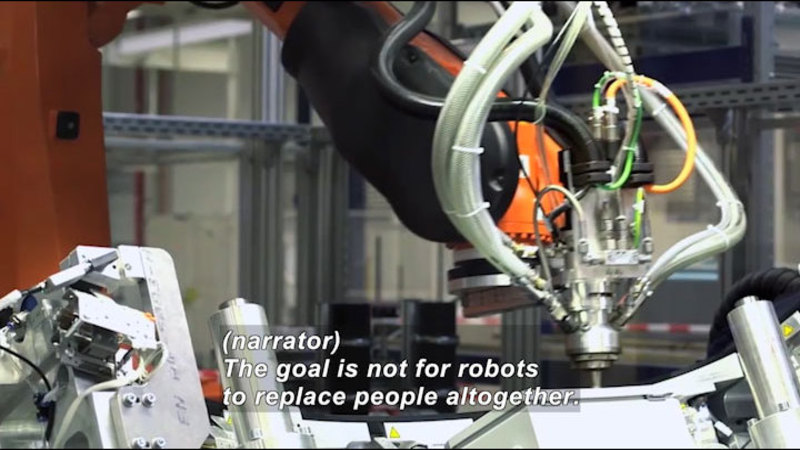 Robotic arm working on an object. Caption: (narrator) The goal is not for robots to replace people altogether.
