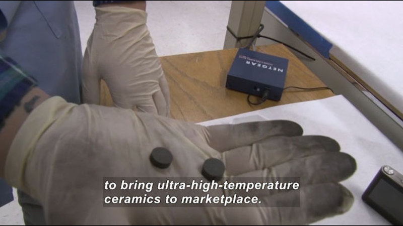 Gloved hand holding two small dark discs. Caption: to bring ultra-high-temperature ceramics to marketplace.