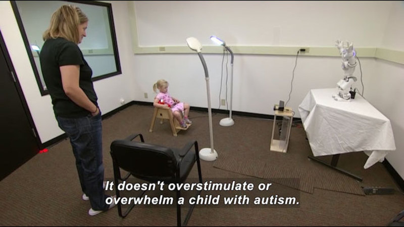 Child sitting in a chair under bright lights looking at a small humanoid robot on a table some distance in front of them. An adult stands by. Caption: It doesn't overstimulate or overwhelm a child with autism.
