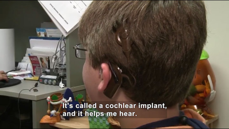 Person with a device attached to their head behind and above their ear and a wire leading to a device that is affixed to the back of their ear. Caption: It's called a cochlear implant, and it helps me hear.