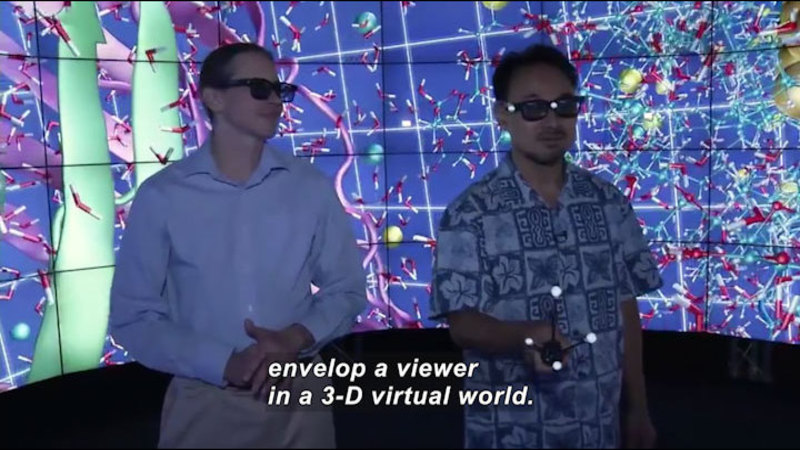 Two people wearing dark glasses in front of a wall of monitors displaying geometric objects. One person holds a device with four glowing orbs on legs in his hand. Caption: envelop a viewer in a 3-D virtual world.