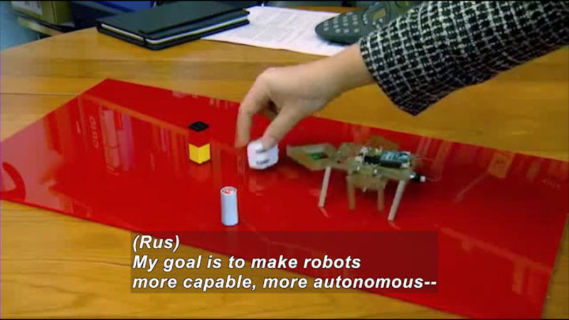Person adjusting objects on a table next to a mechanical object with legs. Caption: (Rus) My goal is to make robots more capable, more autonomous--