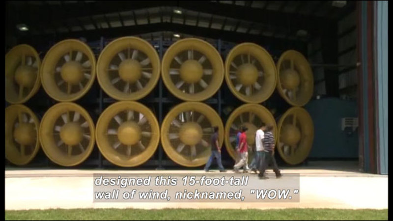 Circular turbines stacked two-high. People walk in front of them. Caption: designed this 15-foot-tall wall of wind, nicknamed, "WOW."