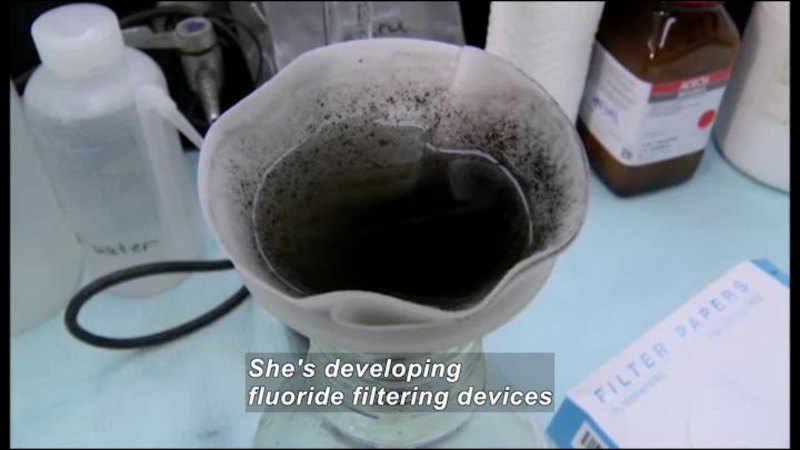 Funnel with filter feeding into a receptacle. The fluid in the filter is dark and the filter is dirty. Caption: She's developing fluoride filtering devices