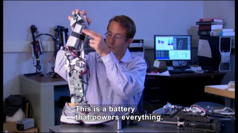 Person working on a robotic leg that is wearing a shoe. Caption: This is a battery that powers everything.