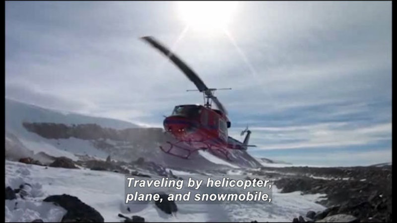 Helicopter hovering over a rocky, snow covered landscape. Caption: Traveling by helicopter, plane, and snowmobile,