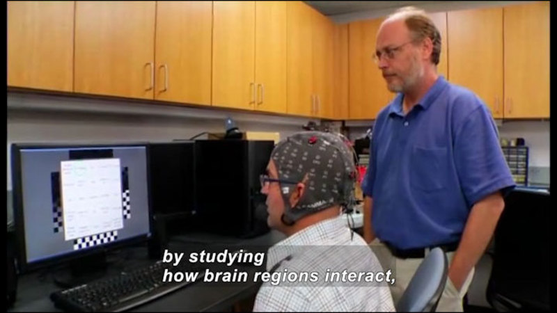 Person wearing a cap with wires and sensors attached while looking at a computer screen. Caption: by studying how brain regions interact,