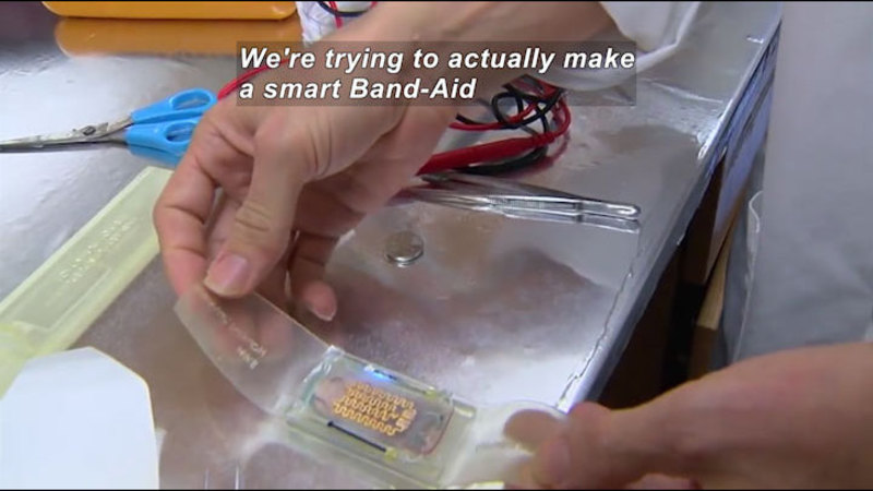 Person in a lab setting with a mostly clear object in their hands. Scissors, wires with leads, and tweezers are on the counter next to them. Caption: We're trying to make a smart Band-Aid