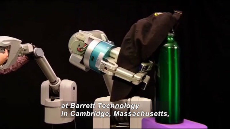 Robotic arm placing a baseball hat on a water bottle. Caption: at Barrett Technology in Cambridge, Massachusetts,