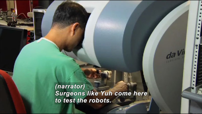 Person in scrubs looking into an enclosed space while manipulating controls with both hands. Caption: (narrator) Surgeons like Yuh come here to test the robots.