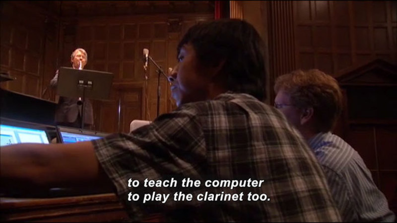 Two people sitting at computer screens while a person on stage plays a wind instrument. Caption: to teach the computer to play the clarinet too.