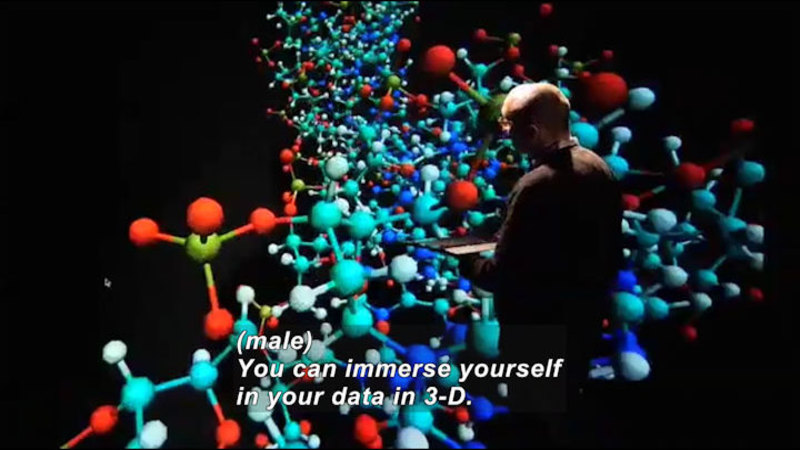Person standing with a touch screen in their hands in front of a screen d9male) displaying 3D representations of the connections between spherical objects. Caption: (male) You can immerse yourself in your data in 3-D.
