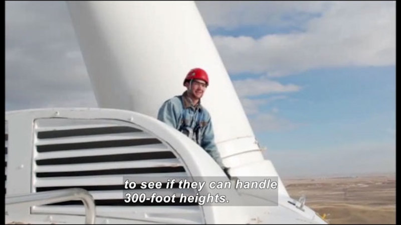 Person standing at the top of a wind turbine. Caption: to see if they can handle 300-foot heights.