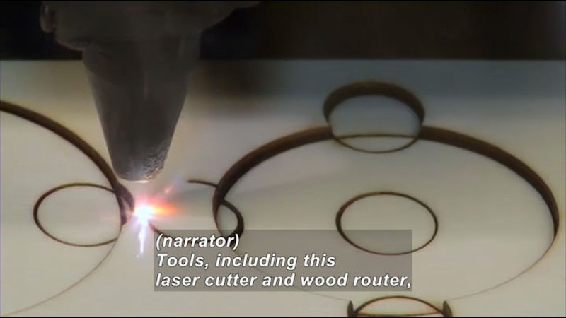 Tool emitting bright light cutting circles into wood. Caption: (narrator) Tools, including this laser cutter and wood router,