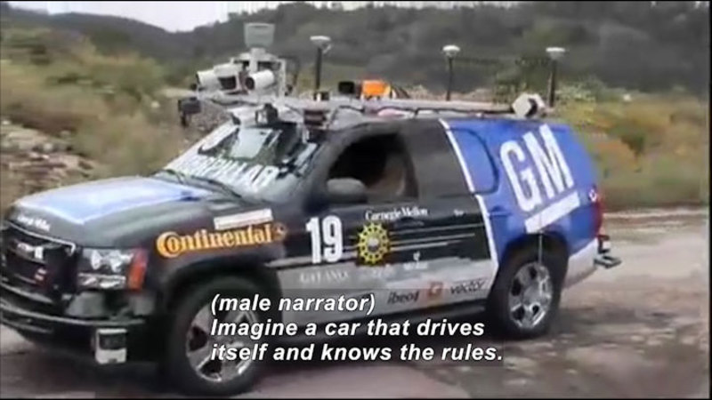 SUV with sensors, cameras, and other equipment attached to the bumper, hood, and roof. Caption: (male narrator) Imagine a car that drives itself and knows the rules.