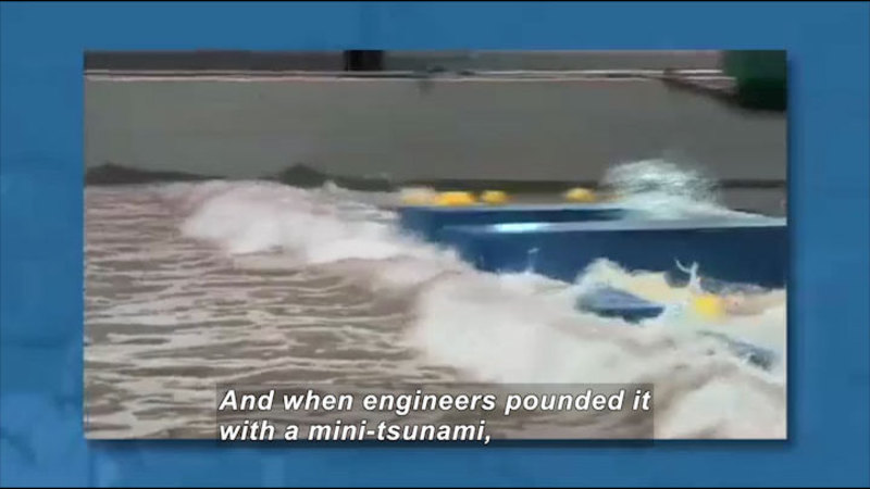 Model building being pounded by waves. Caption: And when engineers pounded it with a mini-tsunami,