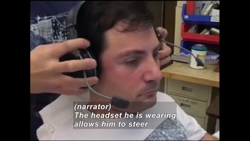Person with a headset being placed on their head. Caption: (narrator) The headset he is wearing allows him to steer