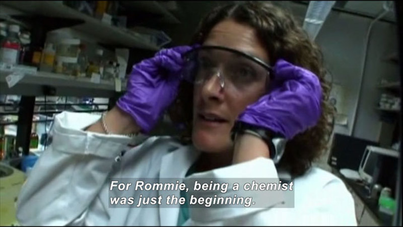Person in a lab coat and gloves putting on protective glasses. Caption: For Rommie, being a chemist was just the beginning.
