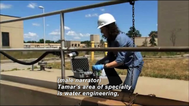 Person wearing a hard hat kneeling next to a metal fence working on something. Caption: (male narrator) Tamar's area of specialty is water engineering,