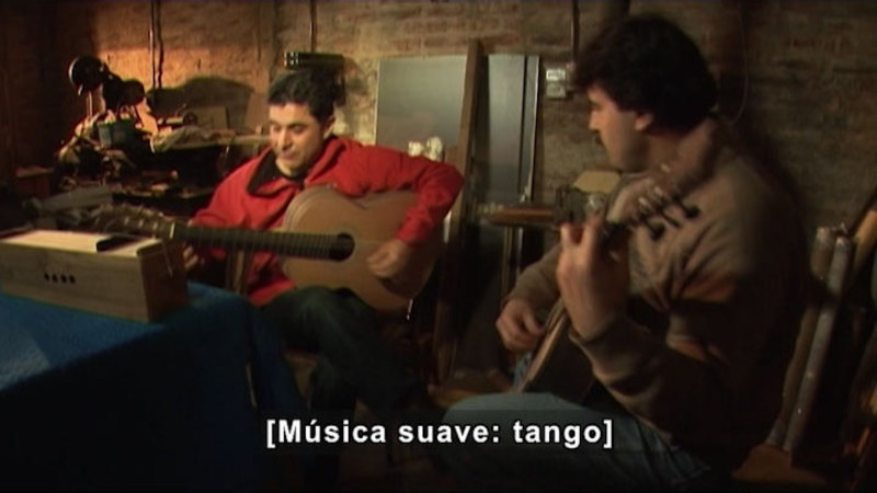 Two people sitting and playing guitars. Spanish captions.