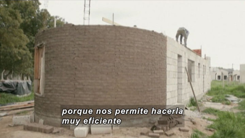 Building with a rounded end under construction. Person working on the roof. Spanish captions.