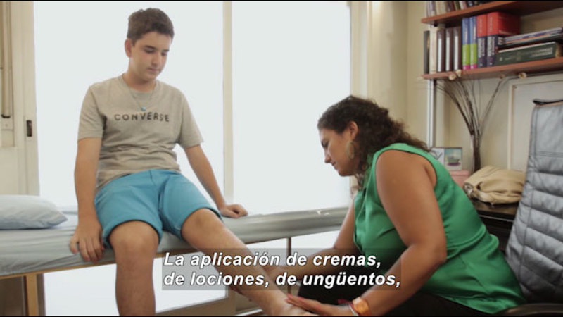 Young person sitting on an examination table with leg extended while someone looks at it. Spanish captions.