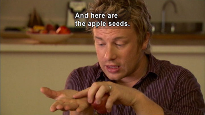 Person holding an apple in one hand and pointing at a small object on the palm of their other hand. Caption: And here are the apple seeds.