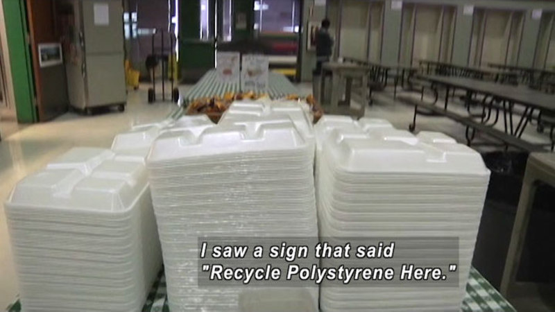 Stacks of disposable meal trays in foreground with a school cafeteria in the background. Caption: I saw a sign that said "Recycle Polystyrene Here."
