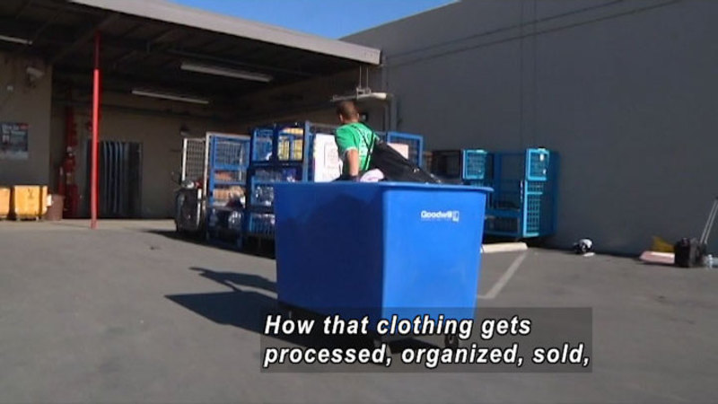 Person pulling a blue bin with "Goodwill" printed on it across a paved loading dock. Caption: How that clothing gets processed, organized, sold,