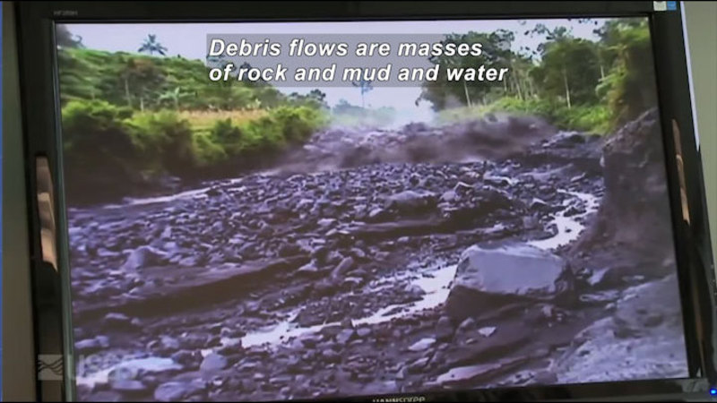 Computer screen showing a dry riverbed with a rushing wall of wet debris. Caption: Debris flows are masses of rock and mud and water