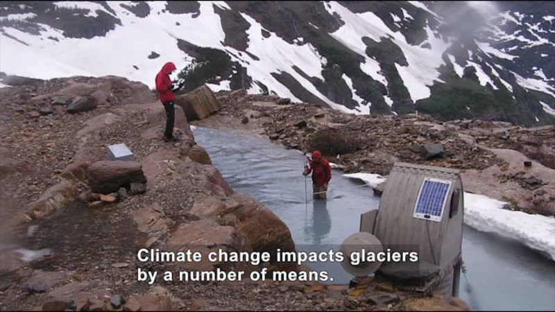 Two people at a glacier. Snow on the mountain in the background, one person standing on a rocky riverbank, the second person in the river with a measurement tool. Caption: Climate change impacts glaciers by a number of means.