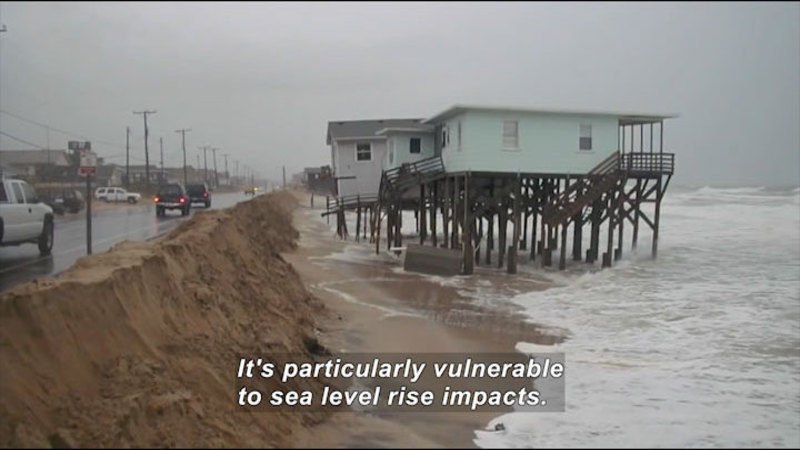 Rough ocean lapping against dirt and sand embankment against a road with vehicles driving on it. Two houses over the water on stilts are starting to fall into the ocean. Caption: It's particularly vulnerable to sea level rise impacts.
