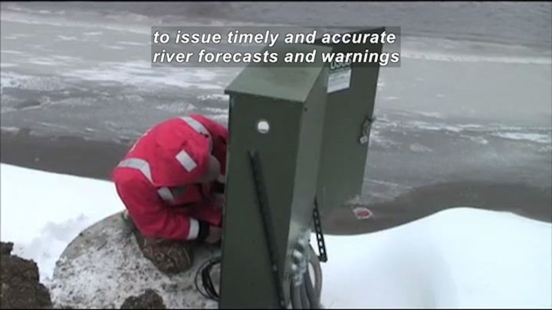 Person crouched next to an electrical box on the bank of a river. Caption: to issue timely and accurate river forecasts and warnings