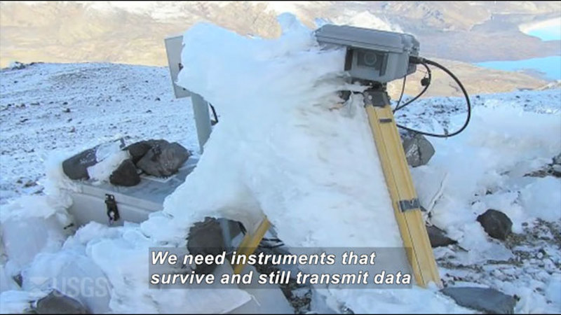 Camera on a tripod next to other equipment, covered in ice and snow. Caption: We need instruments that survive and still transmit data
