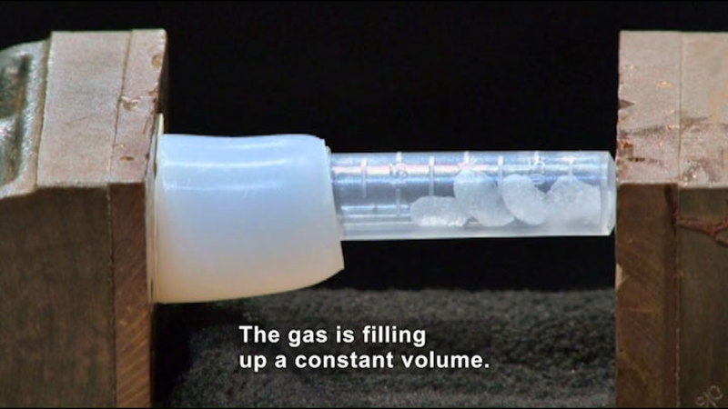 Plastic tube containing clear, pebble-like objects is attached to a larger tube. Caption: The gas is filling up a constant volume.
