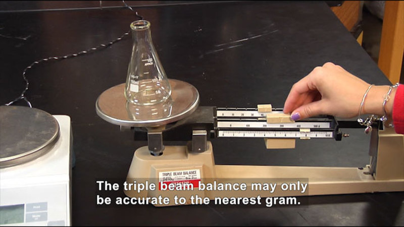 Person weighing a beaker on a triple beam scale. Caption: The triple beam balance may only be accurate to the nearest gram.