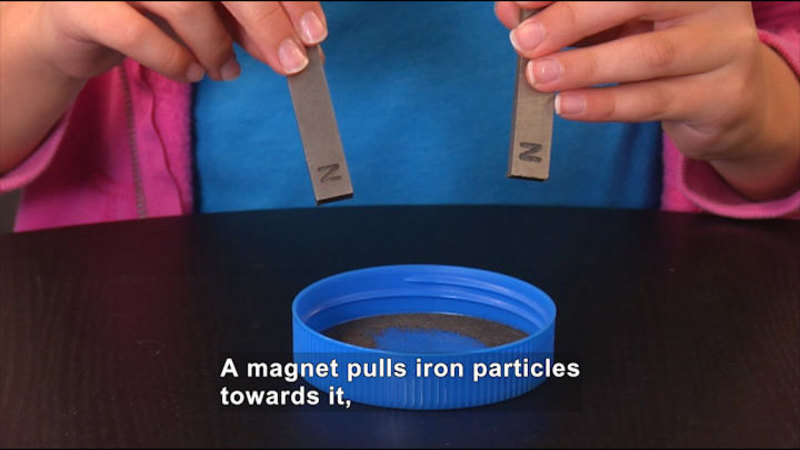 A person holds two magnets over a lid with metal particles in the bottom. Caption: A magnet pulls iron particles towards it,